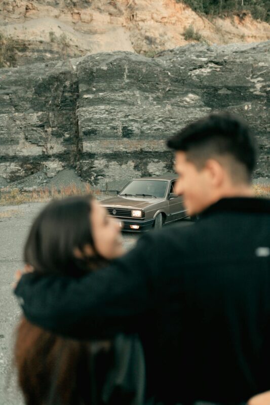 A couple embraces while admiring a classic car parked near rocky cliffs.
